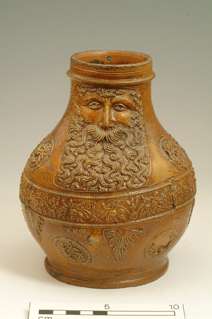 Stoneware jug with pale brown salt glaze. It is decorated with a bearded man's face on the neck, a line of foliage around the waist and leaves, some of which are in circles.