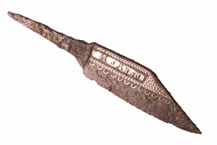 Iron knife. It has inlaid silver decoration of a row of semi-circles, a herringbone pattern and letters of an unreadable word. The handle is missing.