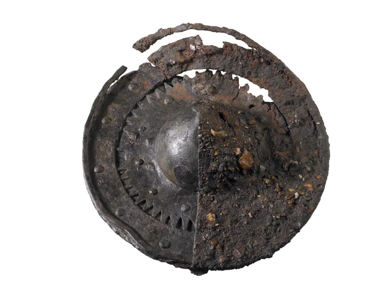 Small, circular shield with a boss in the centre. Half of the shield has been conserved and the other has been left covered in corrosion.