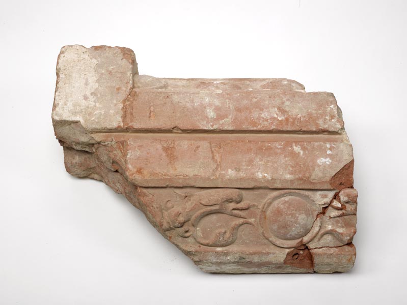 Part of a terracotta moulding in reddish-brown clay, with remains of white plaster over the surface. It is decorated with a leaf pattern along one side.