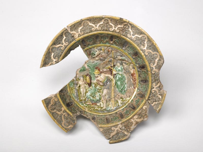 Small, highly decorated and multi-coloured plate. In the centre is a classical scene of the goddess Minerva with Cupid and other figures in an exotic landscape.