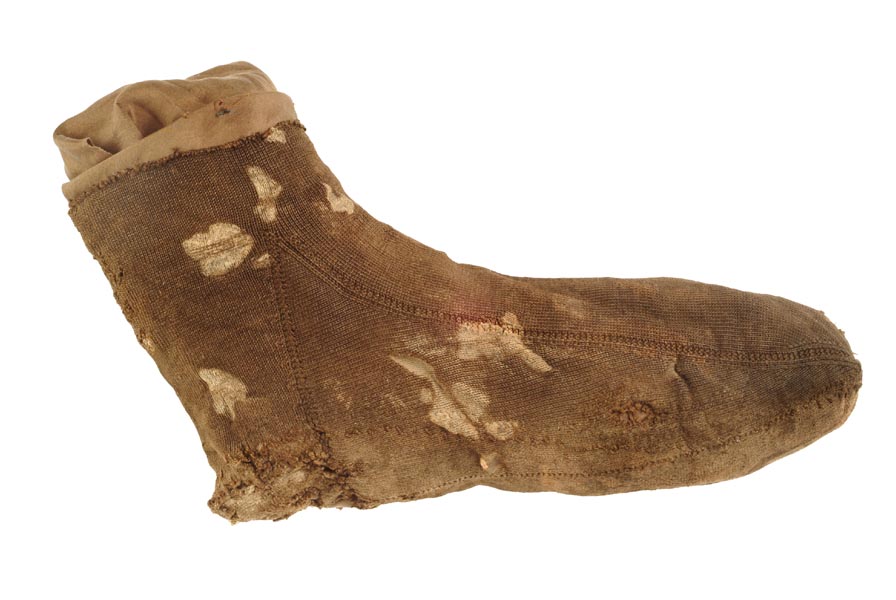 Foot of a silk stocking. The silk is a brown colour, with patches of cream. The knitting is very fine. There is a mended hole in the heel.