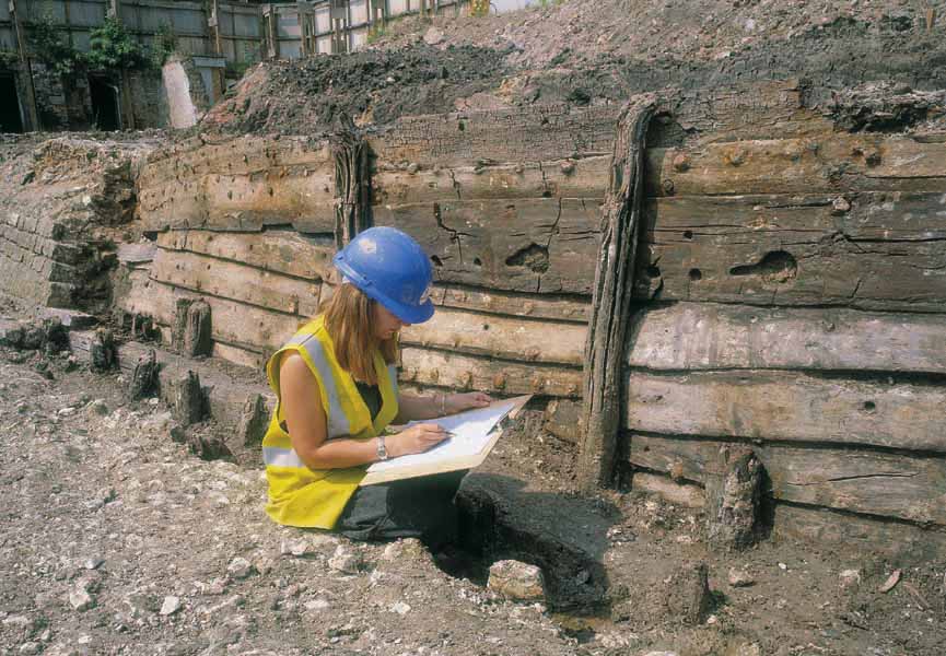 Photograph of the archaeological excavation of the side of a wooden ship. In the foreground is an archaeologist drawing. Behind her are the timbers preserved in the mud.