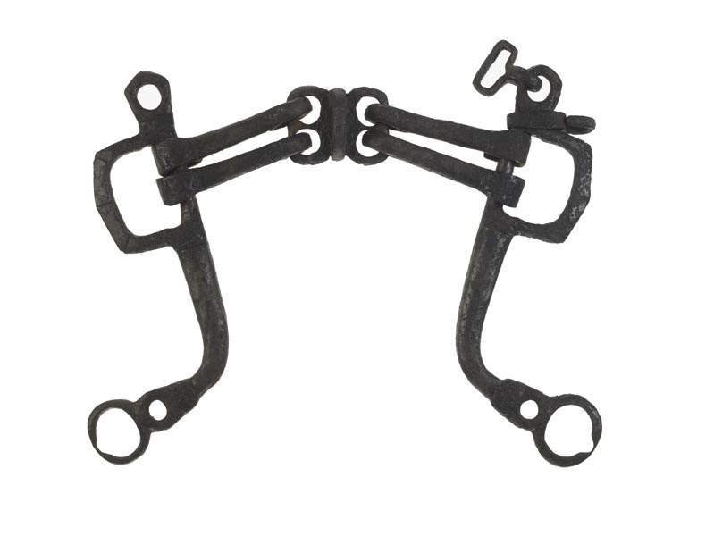 Large iron horse bit with long, L-shaped cheek pieces at each side. At the bottom of these are the loops for the reins, which are nearly worn through with use.